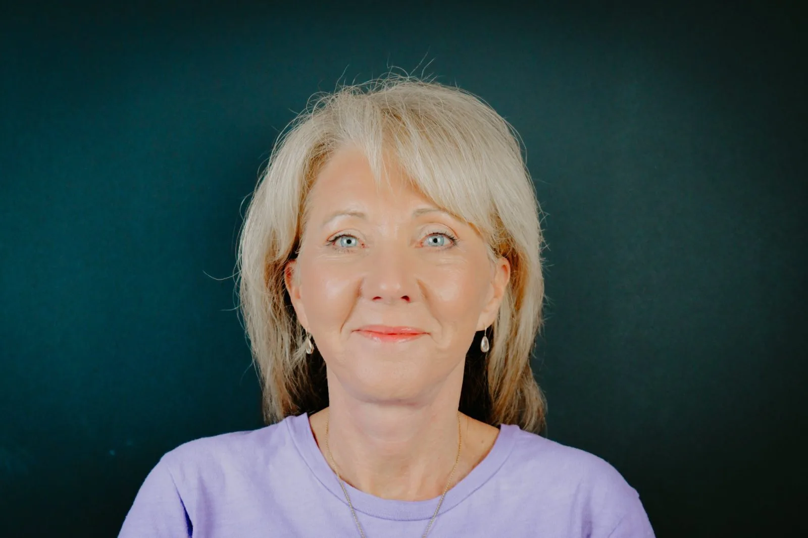 Woman with shoulder-length blonde hair and blue eyes wearing a lavender shirt against a teal background
